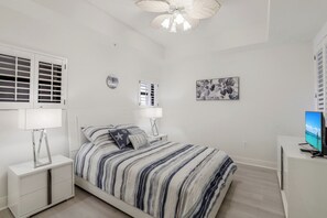 Ein Schlafzimmer mit Bett, Nachttischen, Deckenventilator, Fernseher und einem Fenster mit Rollläden.