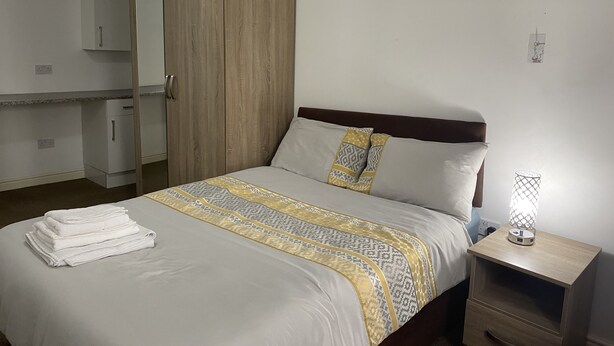 A neatly made bed with a patterned bedspread, a wooden wardrobe, a bedside table with a lamp, and a small cabinet. - Inviting 3-bed Apartment in Stockton-on-tees (Stockton-on-Tees)