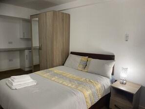 Uma cama bem arrumada com colcha estampada, um guarda-roupa de madeira, uma mesinha de cabeceira com abajur e um pequeno armário.