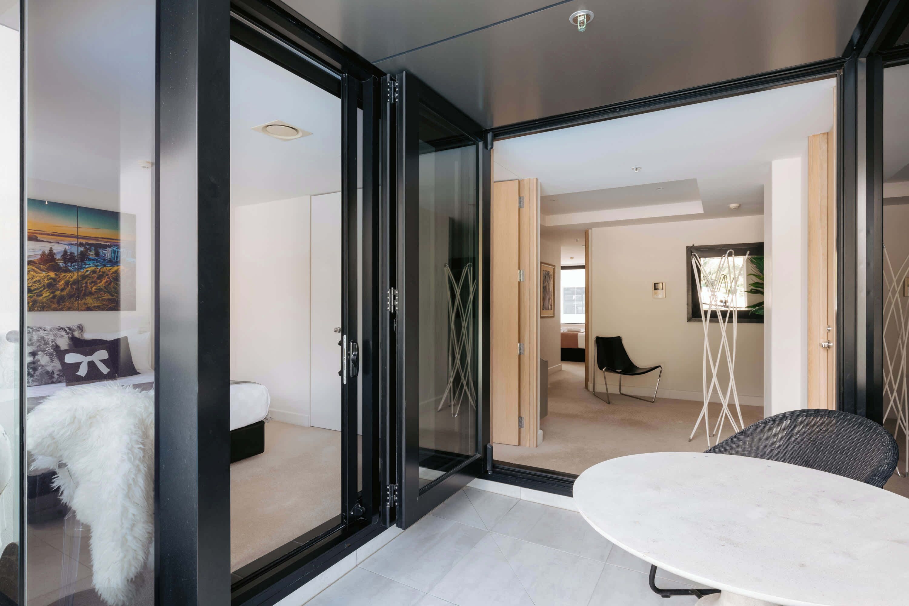 A modern interior with a glass door leading to a bedroom, a white round table, and a wicker chair.