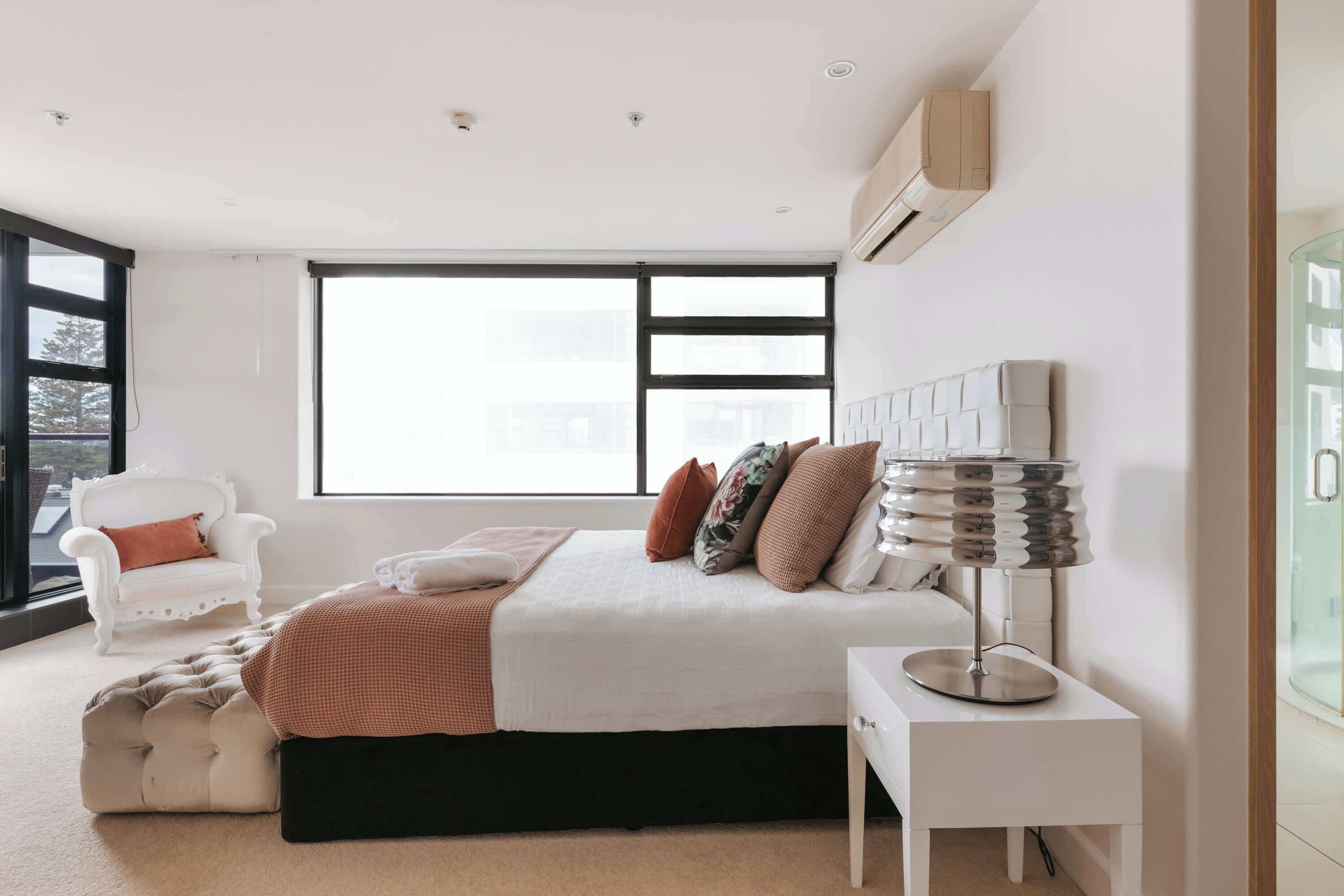 Un dormitorio moderno con un ventanal grande, una cama blanca, una silla blanca y una mesita de noche con lámpara.