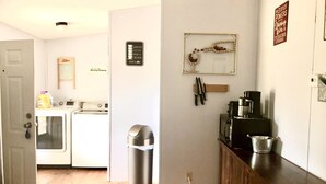 A laundry area with a washing machine, dryer, and a trash can. There is a wooden cabinet with a coffee maker and a kettle on top.
