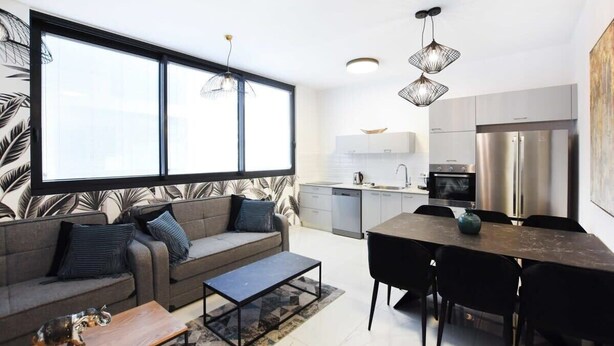 A modern living room with a sofa, coffee table, and dining area with chairs. There is a kitchen in the background with a refrigerator and a sink. - Palm 1BR next to Gazoz Beach by HolyGuest (Tel Aviv-Yafo)