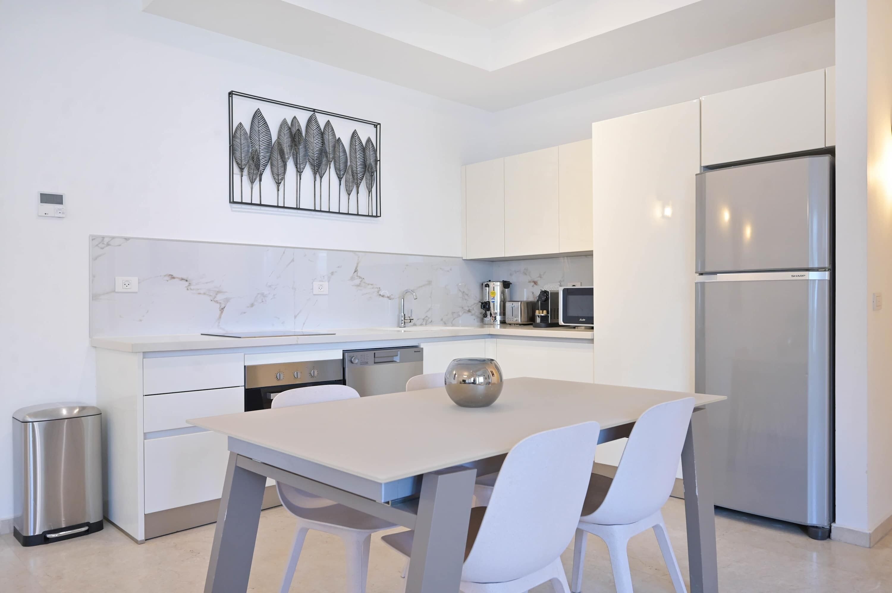 Una cocina moderna con armarios blancos, salpicadero de mármol, refrigerador de acero inoxidable y un comedor con mesa y sillas blancas.