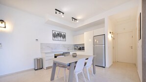 A modern kitchen with white cabinetry, a marble backsplash, and stainless steel appliances. There is a dining table with white chairs and a trash can.