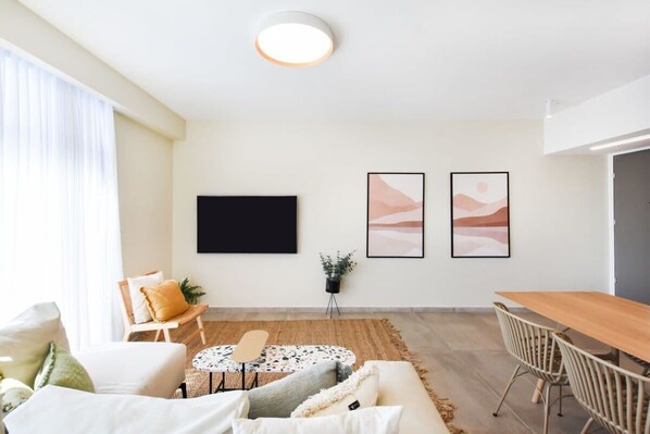 A modern living room with a sofa, a coffee table, a dining table with chairs, a flat-screen TV mounted on the wall, and two framed artworks.