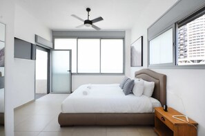 A modern bedroom with a large bed, a ceiling fan, a window with blinds, and a nightstand with a lamp.