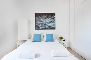 A hotel room with a large bed, white bedding, two blue pillows, a white nightstand with a clock and a small plant, and a painting of a wave on the wall.
