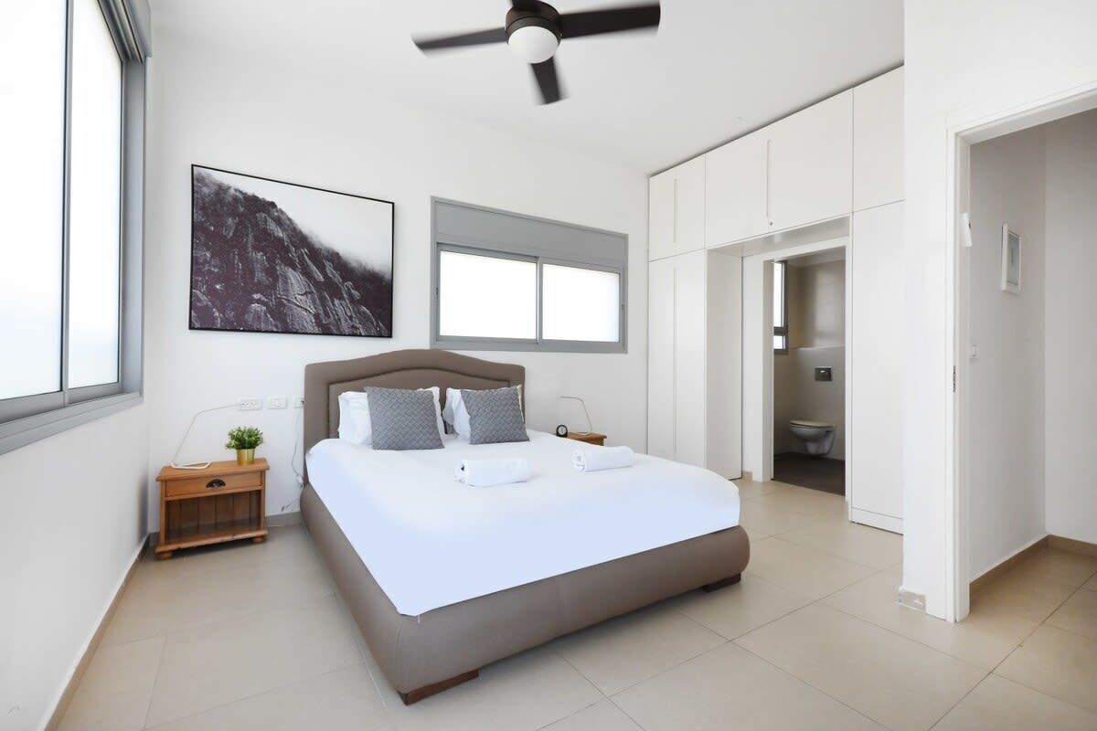 A modern bedroom with a bed, a ceiling fan, a window, a bedside table with a plant, and a bathroom door.
