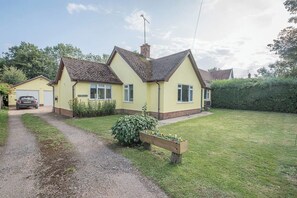 Une maison à deux étages avec une façade jaune, une allée en gravier et un banc en bois dans le jardin.