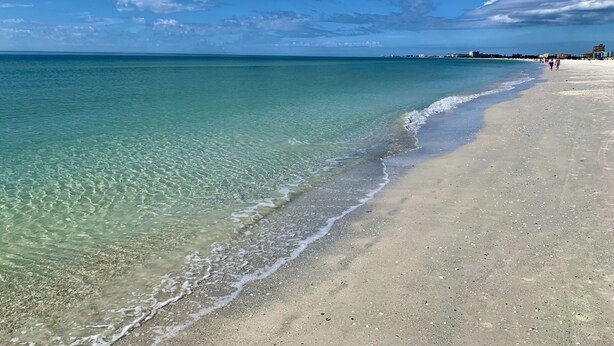 En strand med krystalklart turkis vand, sandet bredde og en fjerntliggende kystlinje med bygninger.