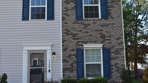 A two-story house with a brick section and a white siding section, featuring a front door, two windows with shutters, and a small front yard with bushes. - Comfortable, Beautiful Townhome in quaint neighborhood w/ patio deck (Henrico)