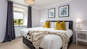 A bedroom with a large bed, two bedside tables, a window with curtains, and two framed pictures on the wall. - Shore View - Donnini Apartments (Prestwick)