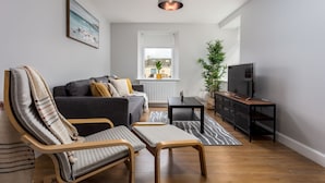 A modern living room with a grey sofa, a wooden chair, a small table, a TV, and a potted plant. - Shore View - Donnini Apartments (Prestwick)
