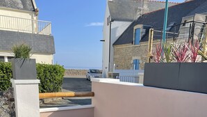 Een terras met een witte tafel en blauwe stoelen, een kamerkleed met een patroon, en uitzicht op zee en gebouwen.