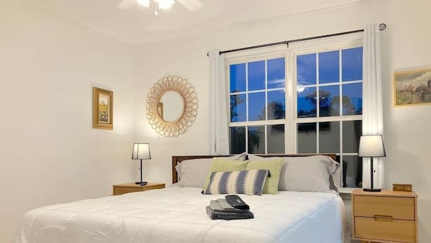Ein Schlafzimmer mit einem großen Bett, zwei Nachttischen, einem Fenster mit Vorhängen, einem Wandspiegel und einem gerahmten Bild.