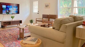 Ein Wohnzimmer mit beiger Couch, Holzstühlen, einem an der Wand befestigten Fernseher, einem gemusterten Teppich und einem Fenster mit Blick auf Grünflächen.