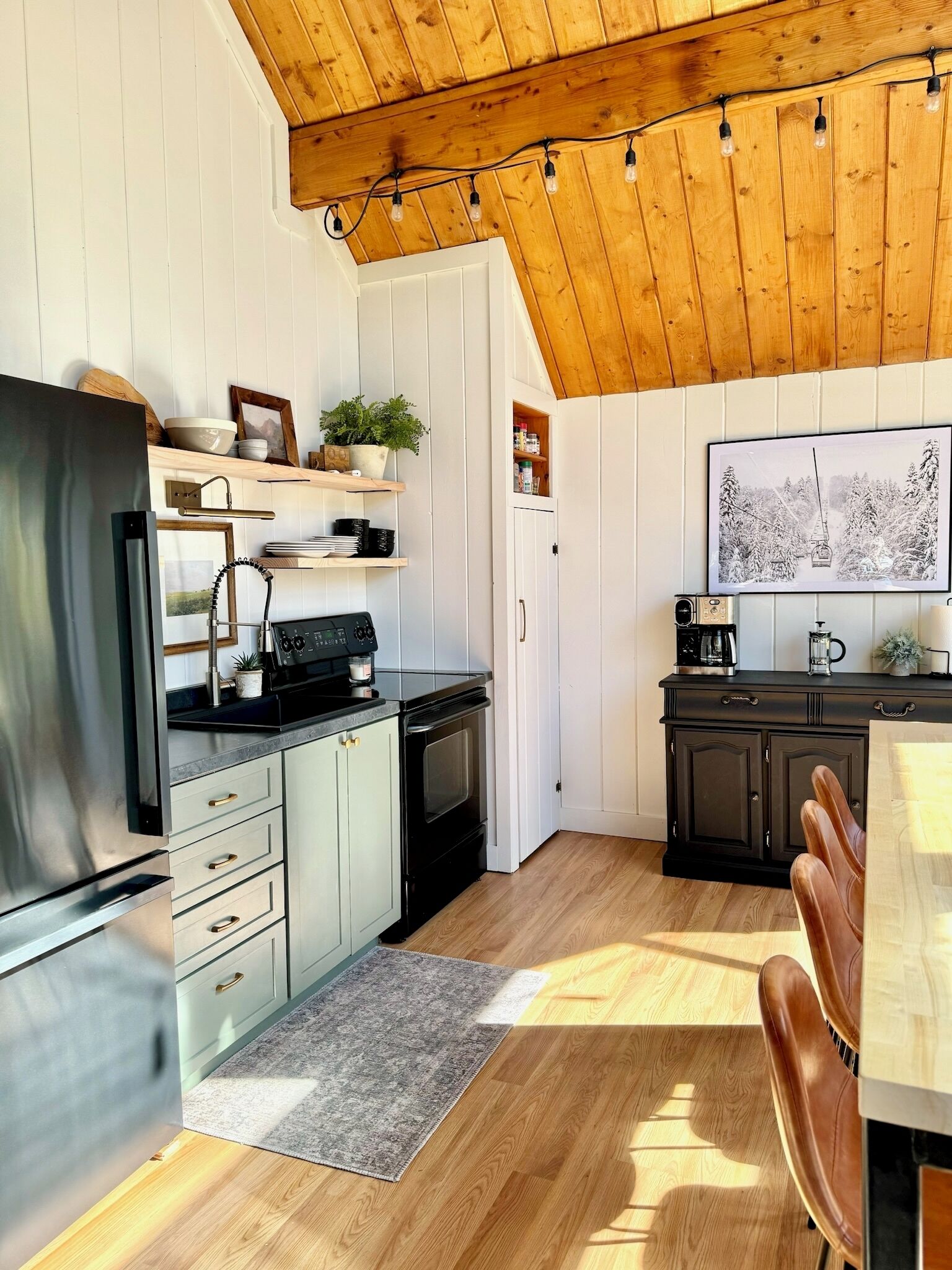 Una cocina moderna con armarios blancos, una estufa negra y un refrigerador de acero inoxidable. Hay estantes de madera con plantas y utensilios de cocina, y un comedor con una silla de cuero marrón.