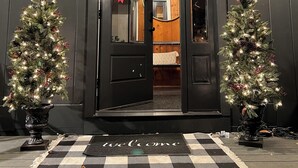 Een donkerhouten deur met een welkomstmat, twee verlichte kerstbomen en een plafondlamp.