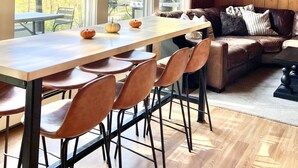 Een ruime kamer met een grote houten eettafel, meerdere stoelen, een bank en uitzicht op herfstbomen door grote ramen.