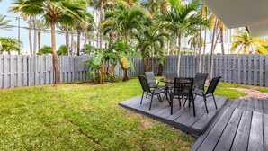 A patio area with a wooden deck, a table with chairs, and a view of palm trees and a fence. - 2 Bedroom 2 Bath Beach 1 min Walk 8 People Keyless Free Parking Flex Check in (Sunny Isles Beach)