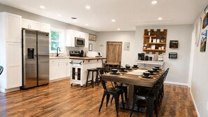A modern kitchen with white cabinets, a stainless steel refrigerator, a wooden dining table with black chairs, and a coffee station. - Modern Family Haven 10 Min to Liberty/ King Bed•Patio•Grill•Firepit•Playset (FOREST)