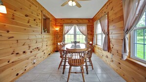 L’intérieur d’un chalet en bois comprend une table à manger et des chaises, un ventilateur de plafond et de grandes fenêtres avec des rideaux.