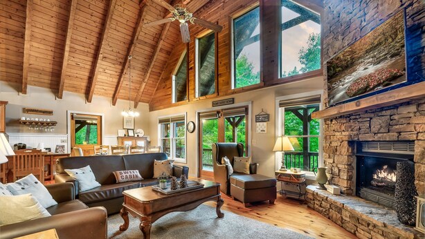 A spacious living room with a stone fireplace, wooden ceiling beams, and large windows offering a view of the outdoors. - Beautiful Views of the Smoky Mountains! Hot Tub-Game Room-3 King Beds-Sleeps 11 (Sevierville)