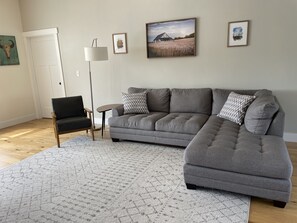 Una sala de estar con un sofá seccional gris, un sillón negro, una lámpara de pie y una alfombra. Hay tres cuadros enmarcados en la pared.