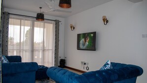 Een woonkamer met twee blauwe banken, een plafondventilator, een televisie en een groot raam met gordijnen.