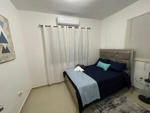 A bedroom with a bed, a bedside table, a lamp, a window with blinds, and an air conditioning unit.