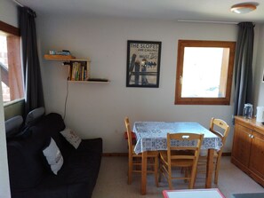 A cozy living room with a dark-colored sofa, a wooden dining table with two chairs, a small bookshelf, and a window with curtains.