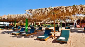 A beachside resort with sun loungers under thatched umbrellas, a sandy beach, and a clear blue sky.
