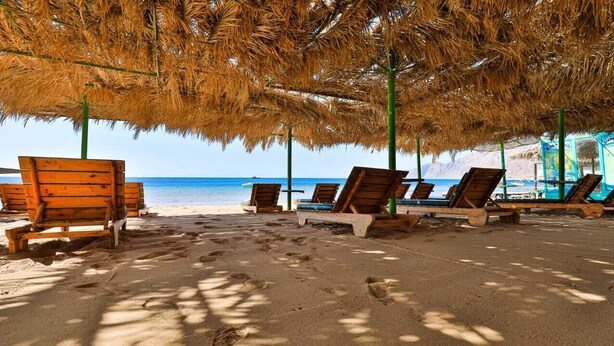 Una playa con sillones de madera bajo un techo de paja, arena y una vista despejada del mar.
