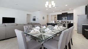 Ein modernes Wohnzimmer mit einem Esstisch aus Glas, gedeckten Plätzen für vier Personen, grauen Sofas und einer Küchenzeile mit dunklen Schränken und einem Edelstahlkühlschrank.