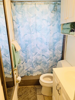 Casa de banho com cortina de chuveiro estampada com folhas, sanita, lavatório e toalheiro com uma toalha verde.