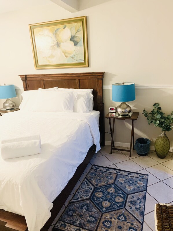 Cama bem arrumada com lençóis brancos, cabeceira de madeira, abajur azul em uma mesa de cabeceira, um relógio, um vaso com folhagem verde e um tapete estampado no chão.