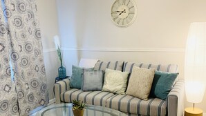Una sala de estar con una mesa de centro de vidrio, un sofá a rayas, almohadones decorativos, un reloj de pared y un ventilador de techo.