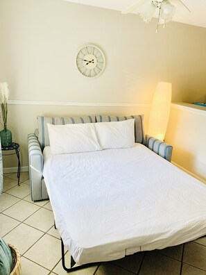 Uma cama bem arrumada com edredom branco e travesseiros, um banquinho estofado listrado, um relógio de parede e um abajur.