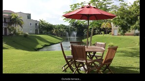 Terraço ao ar livre com mesa e cadeiras, um guarda-sol vermelho e uma fonte ao fundo.