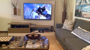 Uma sala de estar com televisão de tela plana na parede, uma mesa de centro com um tabuleiro de xadrez e um sofá com almofadas decorativas.