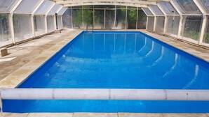 Una piscina coperta rettangolare con acqua cristallina e blu, circondata da un pavimento piastrellato e racchiusa in una struttura di vetro con tetto curvo.