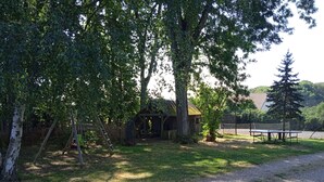 Un sentiero sterrato conduce a una struttura in legno con tetto di paglia, circondata da alberi e un campo da tennis sullo sfondo.