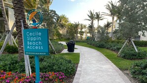 A sign for Cecile Sippin Beach Access in a landscaped area with a paved walkway, palm trees, and flowering plants. - 2 Bedroom 2 Bath Beach 1 min Walk 8 People Keyless Free Parking Flex Check in (Sunny Isles Beach)