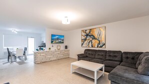 A modern living room with a grey sofa, a white coffee table, and a dining area with a white table and chairs. There is a kitchen area with a stove and a large painting on the wall. - 2 Bedroom 2 Bath Beach 1 min Walk 8 People Keyless Free Parking Flex Check in (Sunny Isles Beach)
