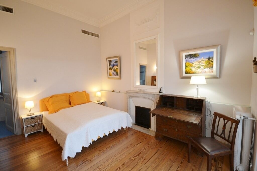 A bedroom with a bed, bedside tables, a desk, a chair, a fireplace, and two framed pictures on the wall.