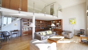 Un'ampia zona giorno con soffitto alto, scaffali in legno, un divano grigio e una scala che conduce a un livello superiore.