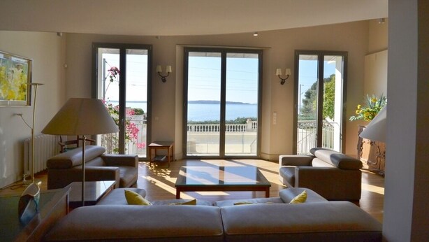 Un ampio soggiorno con un divano grande, un tavolino e due poltrone. Due ampie porte scorrevoli in vetro conducono a un balcone con vista sul mare.