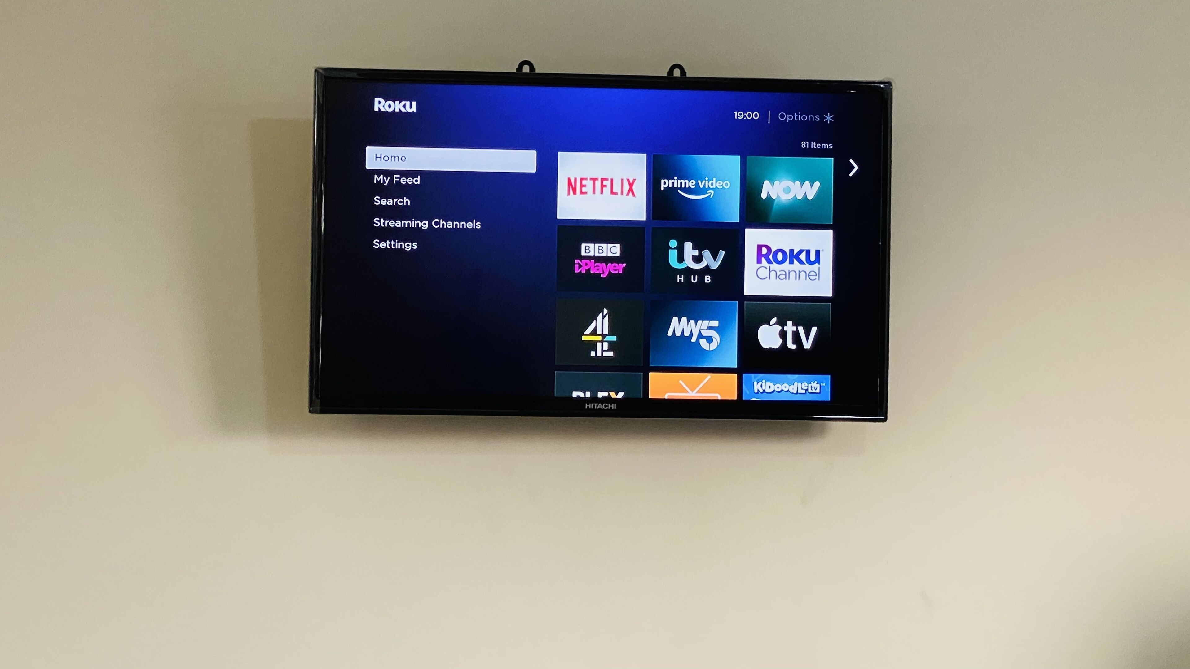 A flat-screen TV mounted on a wall displaying a Roku interface with various streaming channels.