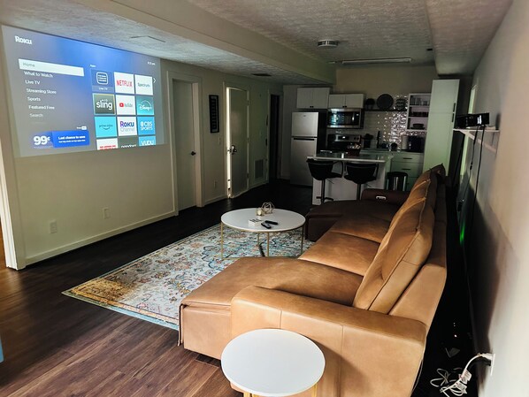 A living room with a brown leather sofa, a white coffee table, and a large screen displaying a streaming service interface.